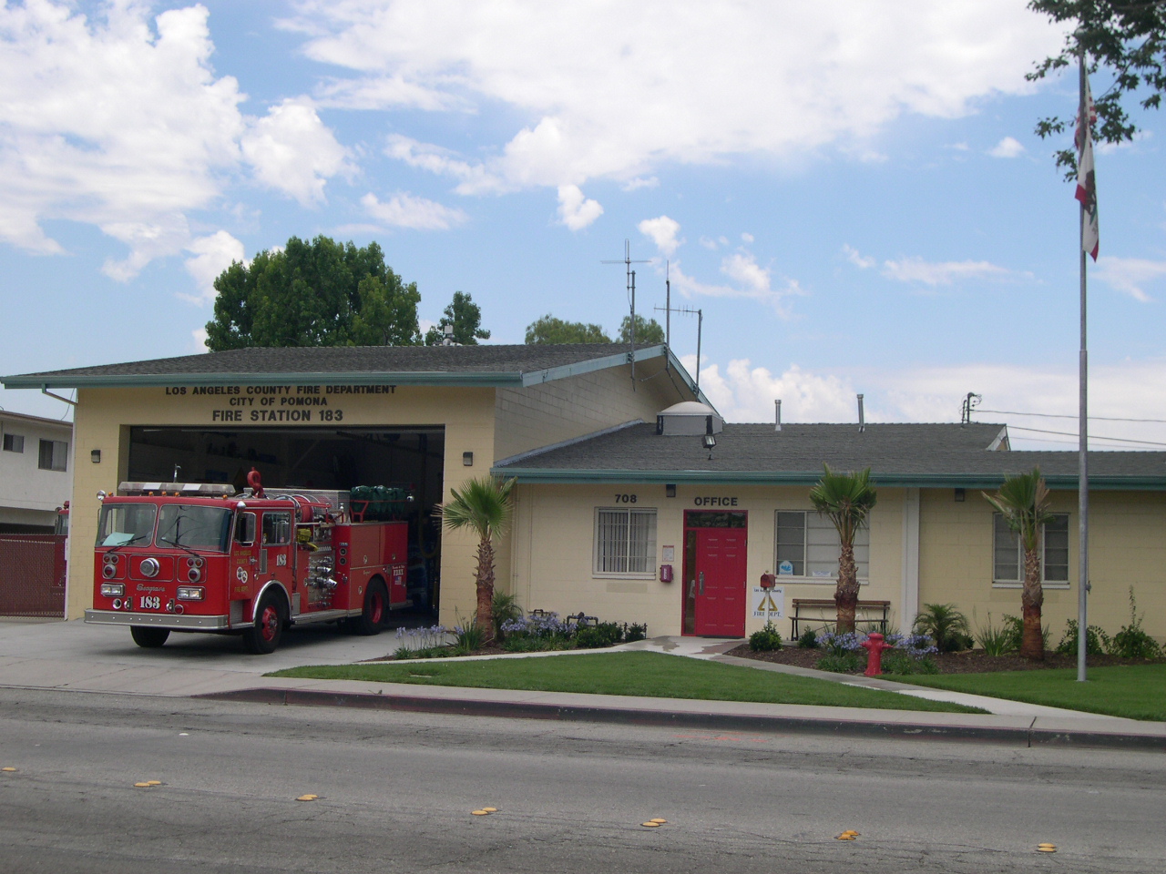 Firetruck 183 parked inside of its station. Station is yellow and has a bright red door. Surrounded by clear skies and the California flag
